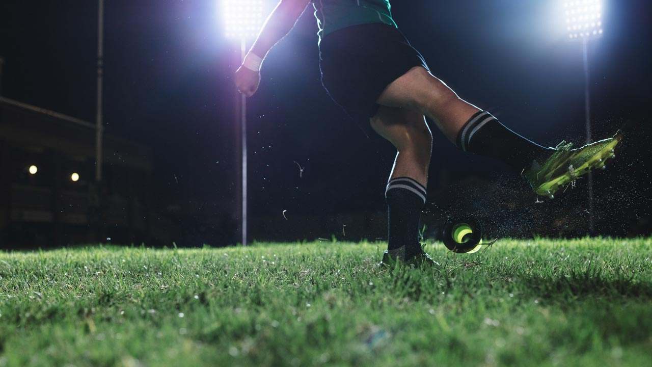 rugby player kicking a ball on pitch at night with spotlight on