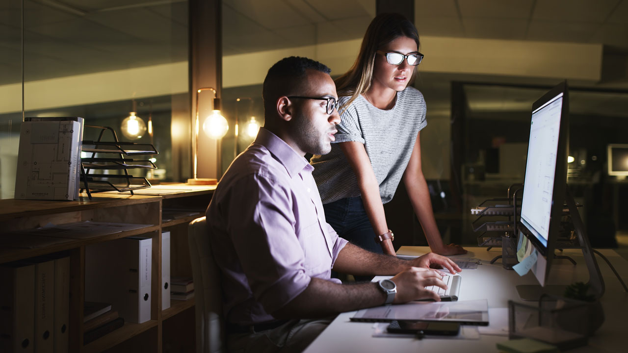 Male seated at desk and female standing, both looking at a computer screen